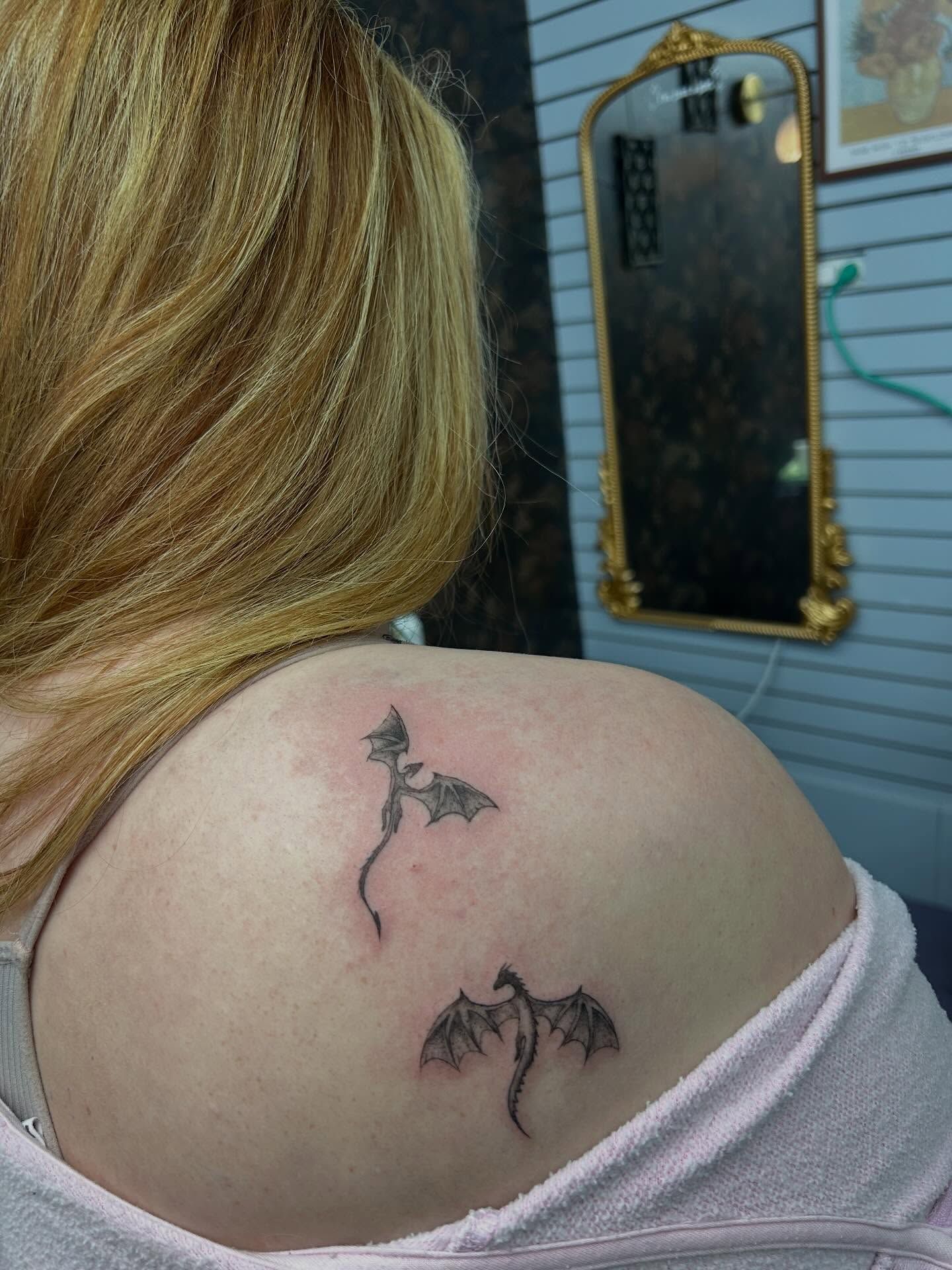 Two small black-and-grey dragons in flight across a shoulder blade