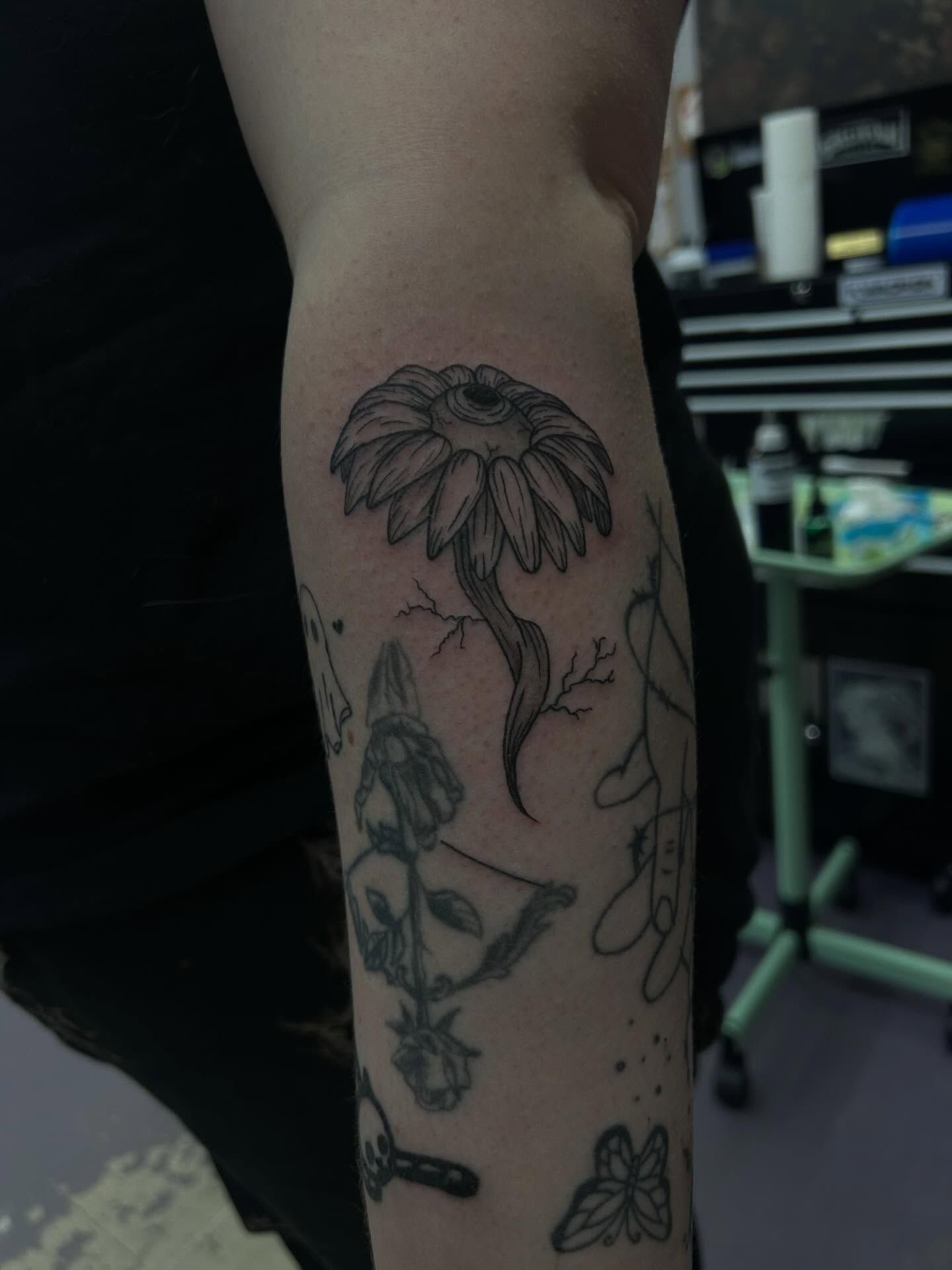 A daisy with a human eye at its centre, in fine black linework on a forearm