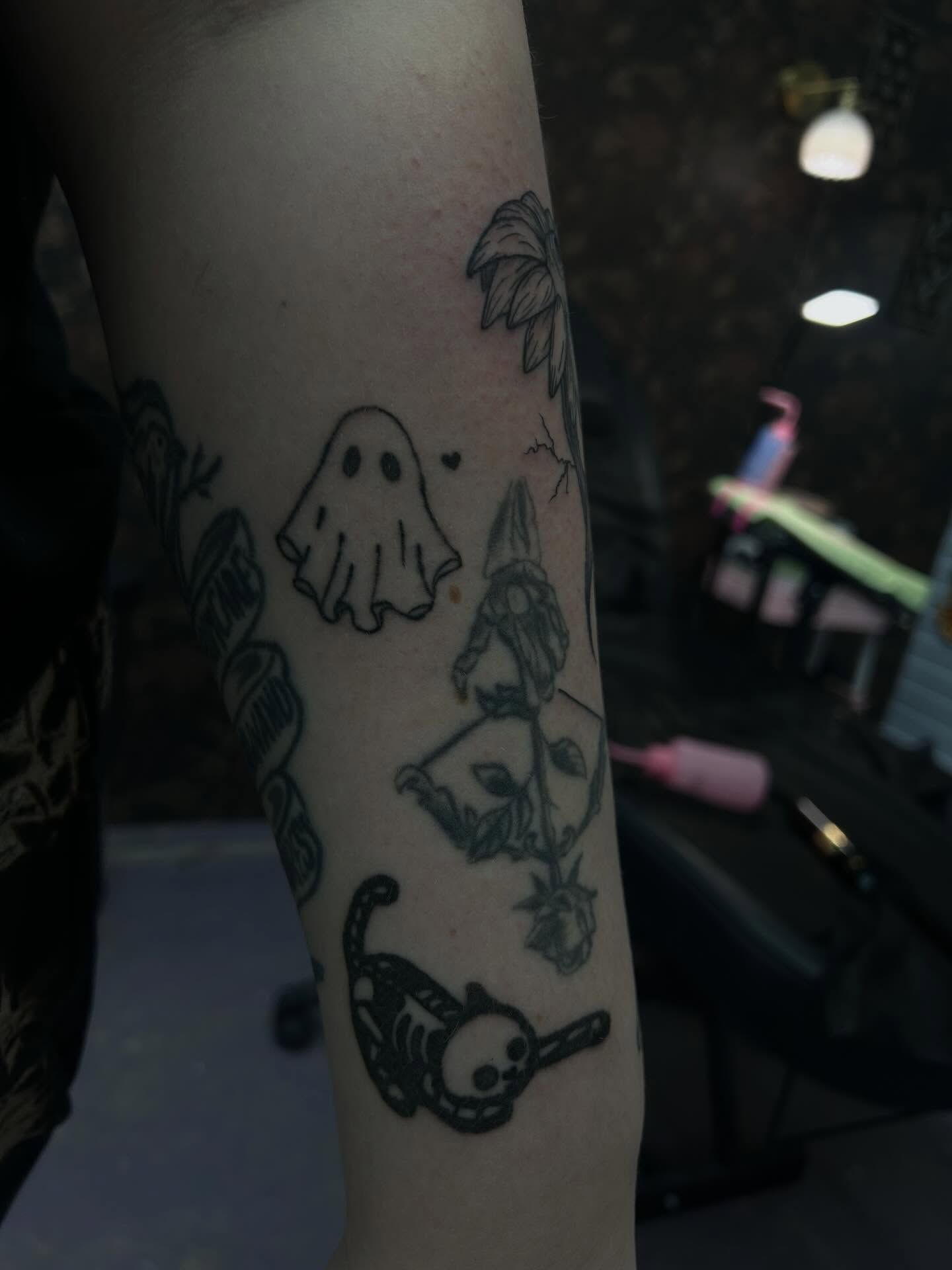 A small outlined ghost with a tiny heart beside it, on an upper arm among other fine-line pieces
