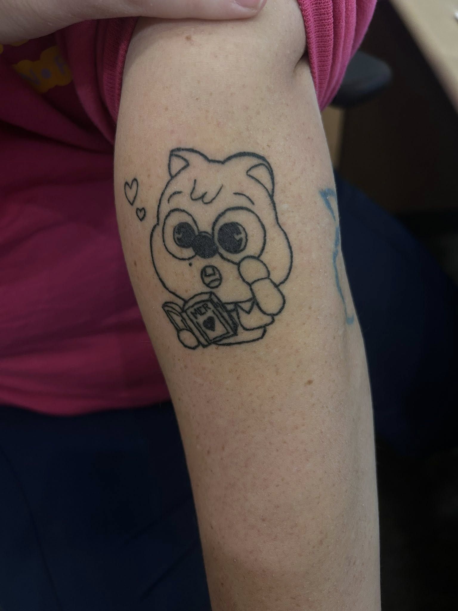 A wide-eyed cartoon cat reading a book, with small hearts floating beside it, on an upper arm