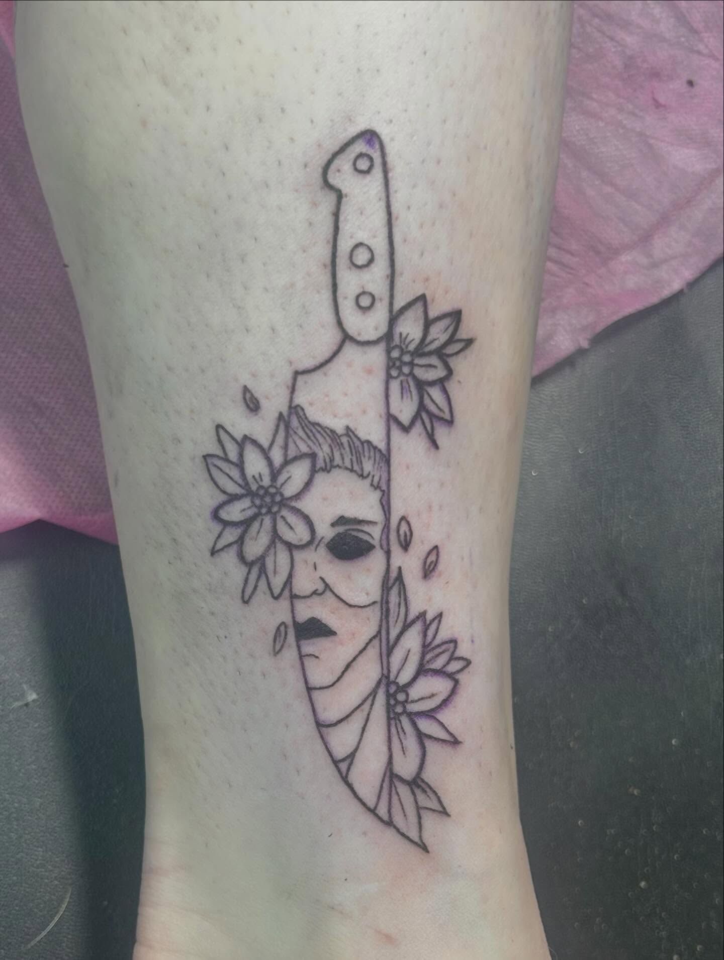 A Michael Myers mask merged into the blade of a chef's knife, surrounded by flowers, on a lower leg