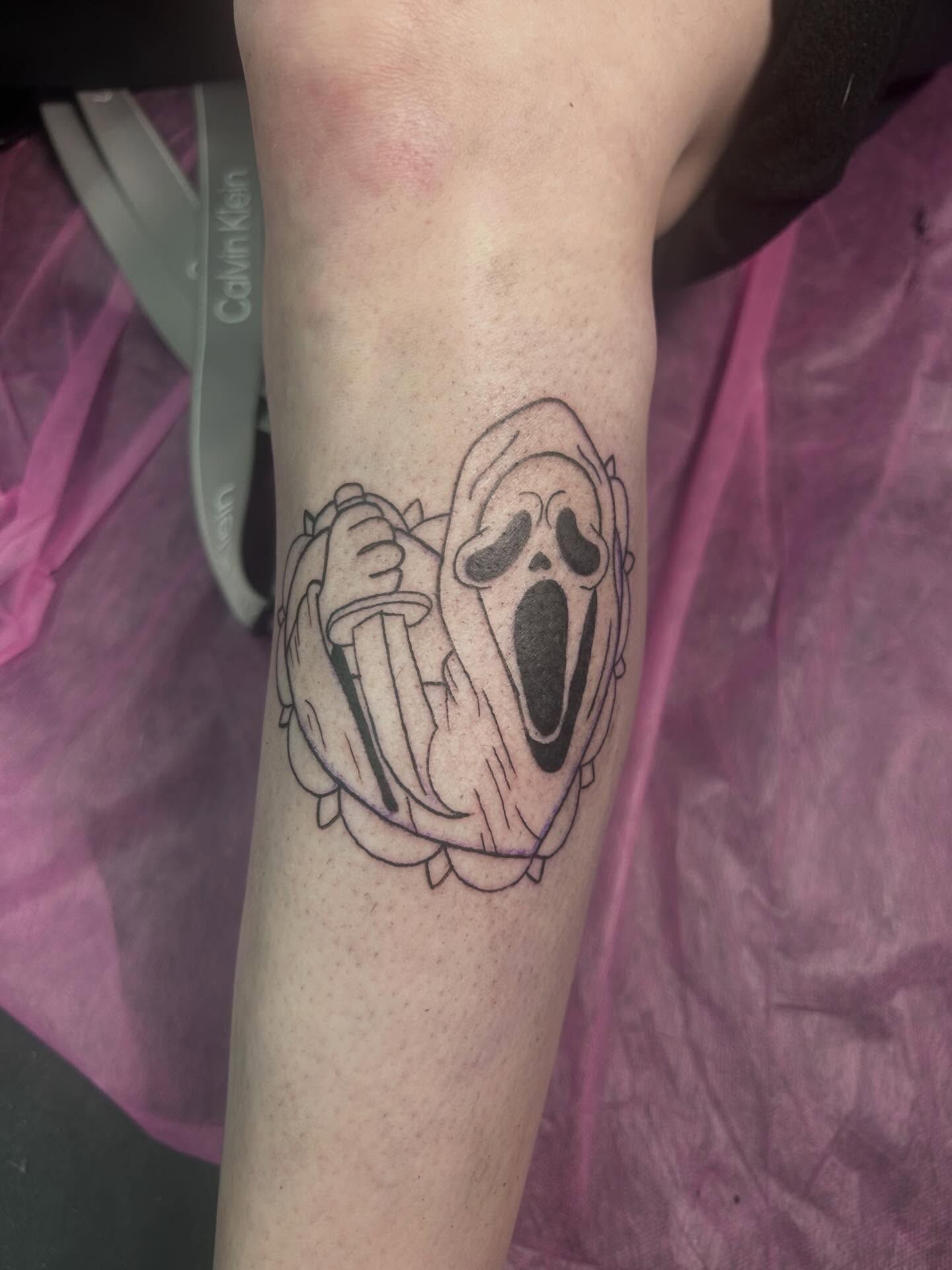 The Ghostface mask holding a knife, framed in a scalloped border, on a forearm